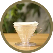V60 Circle 170px A circular image of a clear Hario V60 with a filter paper inside of it. The V60 is sitting on a wooden table.
